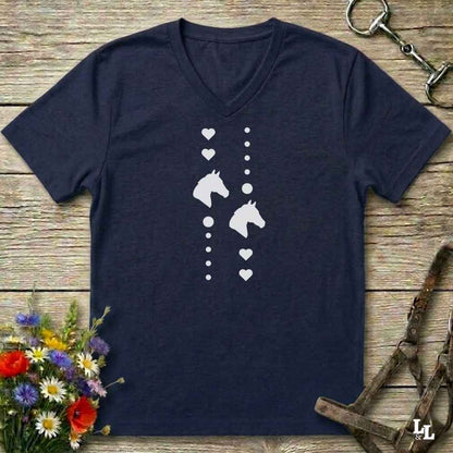 A navy blue V-neck t-shirt with a white graphic print of horse heads, hearts, and dots, laid flat on a rustic wooden background with equestrian accessories and flowers.