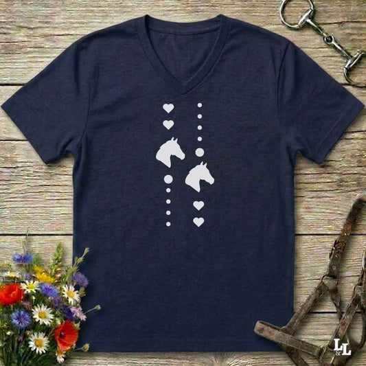 A navy blue V-neck t-shirt with a white graphic print of horse heads, hearts, and dots, laid flat on a rustic wooden background with equestrian accessories and flowers.