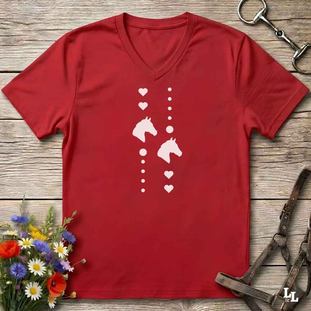 A red V-neck t-shirt with a white graphic print of horse heads and hearts, laid flat on a rustic wooden background with equestrian accessories and flowers.