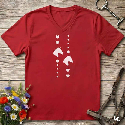 A red V-neck t-shirt with a white graphic print of horse heads and hearts, laid flat on a rustic wooden background with equestrian accessories and flowers.