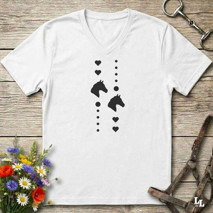 A white V-neck t-shirt with a black graphic print of horse heads and hearts, laid flat on a rustic wooden background with equestrian accessories and flowers.