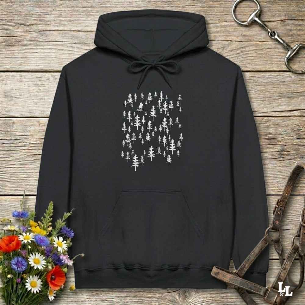 A black pullover hoodie with a white pine tree graphic on the chest, laid flat on a rustic wooden surface with flowers and leather straps as props.