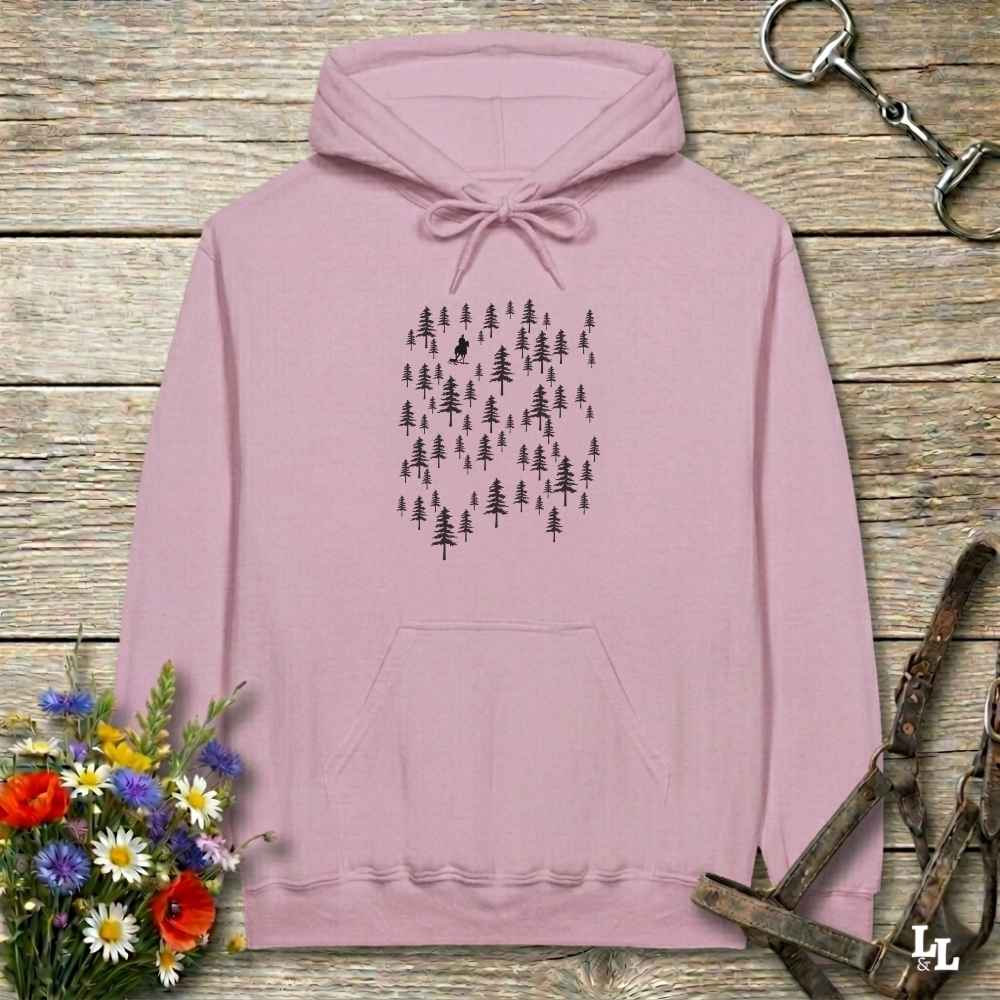 A dusty pink pullover hoodie with a black forest tree print on the chest, laid flat on a rustic wooden background with decorative flowers and leather tack.