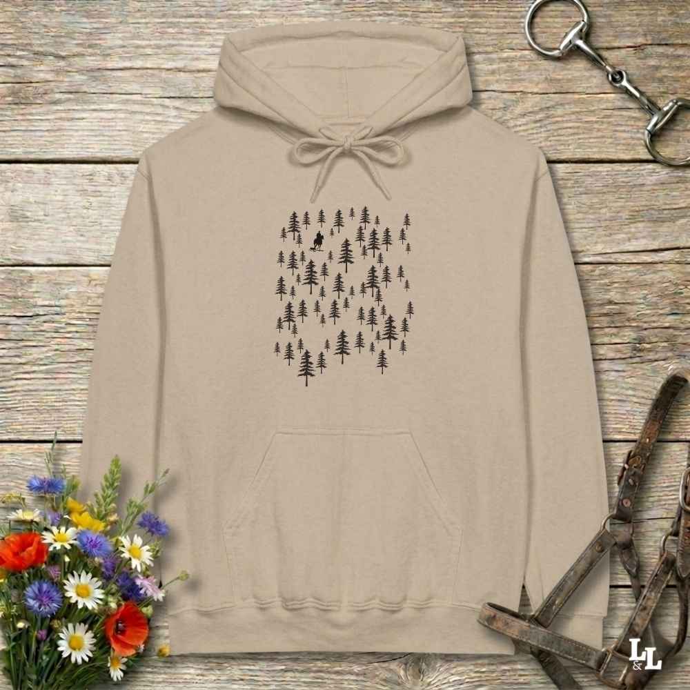 A beige pullover hoodie with a black pine tree forest graphic on the chest, laid flat on a rustic wooden surface with flowers and horse tack props.