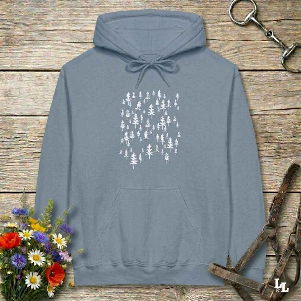 A slate blue pullover hoodie with a white pine tree graphic on the chest, laid flat on a rustic wooden background with flowers and leather tack props.