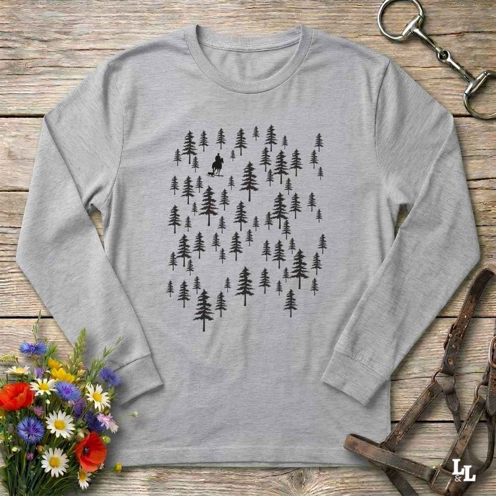 A light gray long-sleeve t-shirt with a black graphic of pine trees and a small mountain biker silhouette, laid flat on a rustic wooden surface with flowers and horse tack props.