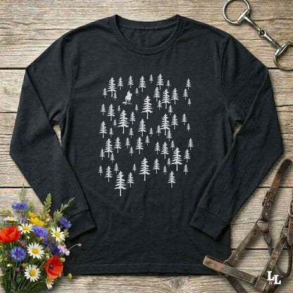A black long-sleeve t-shirt with a white graphic of pine trees, laid flat on a rustic wooden surface with decorative flowers and leather tack around it.