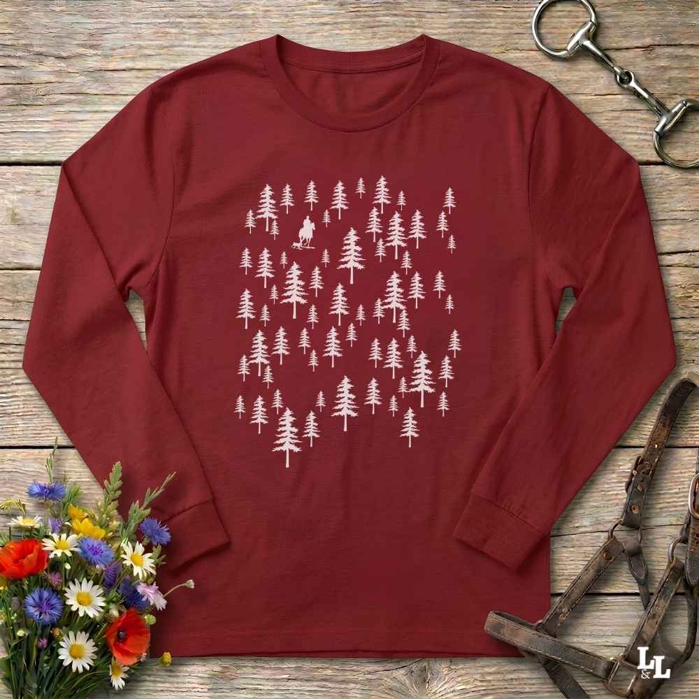 A long-sleeve crewneck t-shirt in maroon red, featuring a white graphic print of various pine tree silhouettes. The shirt is laid flat on a rustic wooden surface, styled with a small bouquet of colorful wildflowers on the bottom left and horse tack (reins and bridle) on the bottom right.