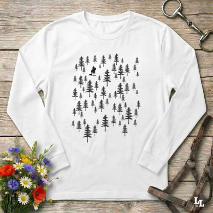 A white long-sleeve crewneck shirt laid flat on a rustic wooden surface, featuring a black graphic of pine trees with a small dog silhouette in the center. The shirt is styled with a bouquet of colorful wildflowers, a horse bit, and leather tack.