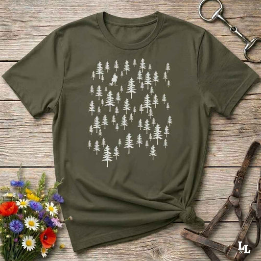 An olive green short-sleeve t-shirt with a white graphic of pine trees, styled in a flat-lay on a rustic wooden background with a horse bit, leather halter, and a small bouquet of wildflowers.