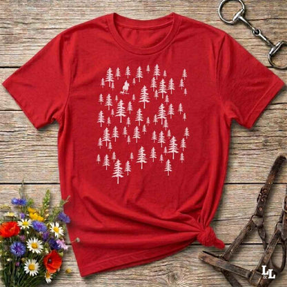 A red short-sleeve t-shirt with a white graphic of pine trees, styled in a flat-lay on a rustic wooden background with equestrian accessories and flowers.