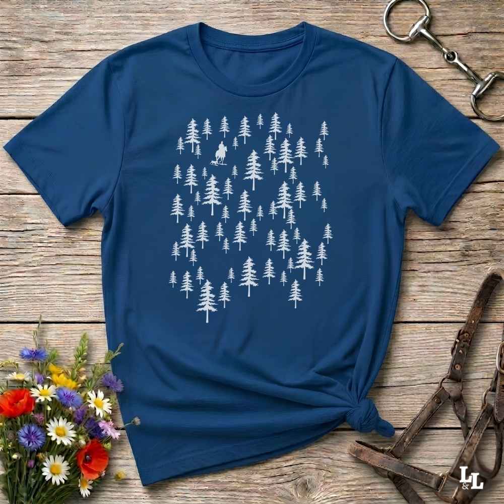 A navy blue short-sleeve t-shirt with a white forest tree pattern, styled in a flat-lay on a rustic wooden background with a horse bit, leather halter, and wildflowers.