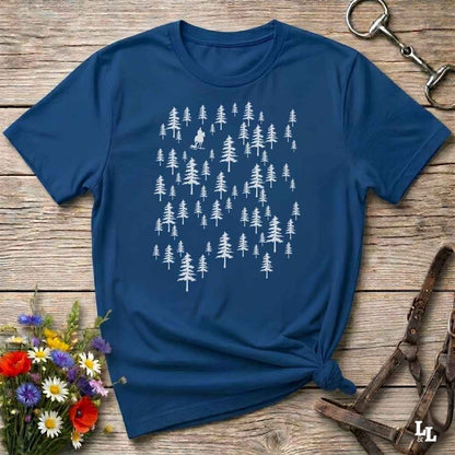 A navy blue short-sleeve t-shirt with a white forest tree pattern, styled in a flat-lay on a rustic wooden background with a horse bit, leather halter, and wildflowers.