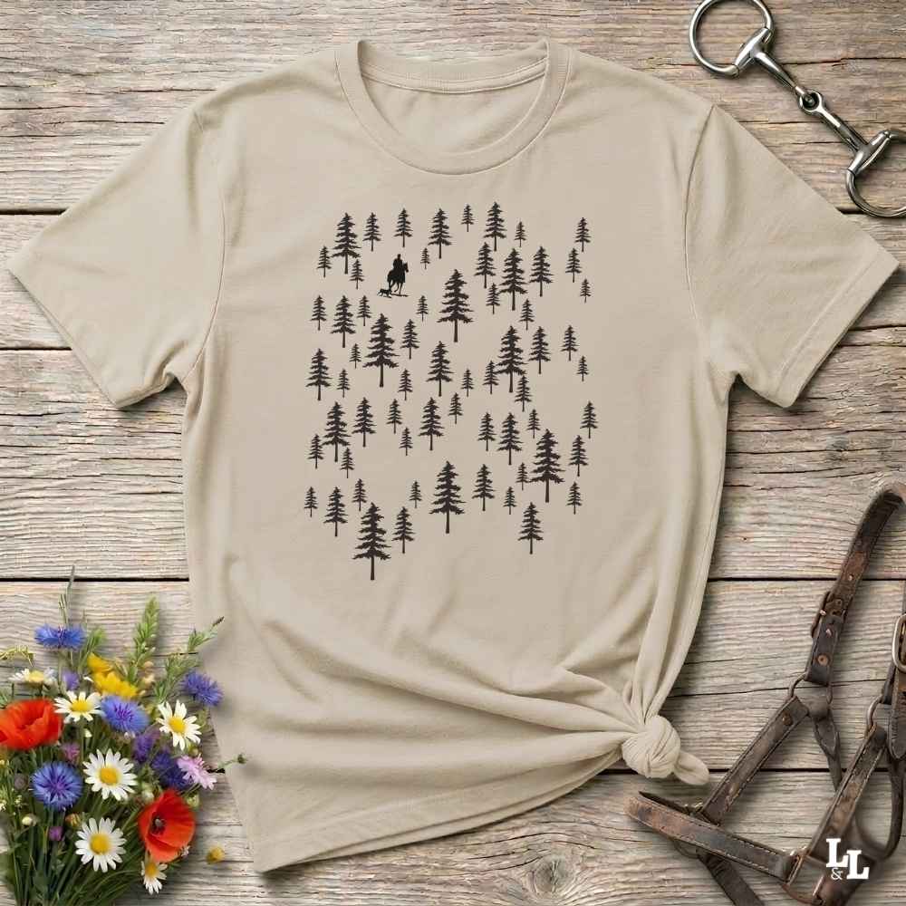 A beige short-sleeve t-shirt with a black forest tree graphic, styled in a flat-lay on a rustic wooden background with a horse bit, leather straps, and a small bouquet of wildflowers.