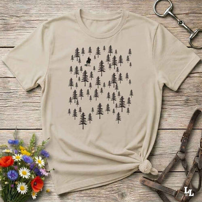 A beige short-sleeve t-shirt with a black forest tree graphic, styled in a flat-lay on a rustic wooden background with a horse bit, leather straps, and a small bouquet of wildflowers.
