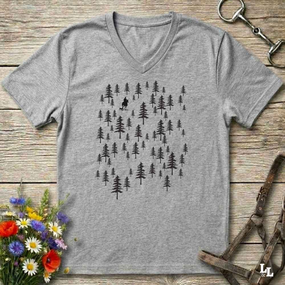 A heather gray V-neck t-shirt with a black graphic of pine trees and a small cowboy riding silhouette, laid flat on a rustic wooden background with flowers and leather gear props.