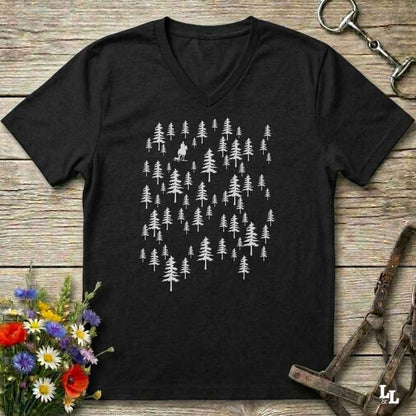 A black V-neck t-shirt with a white graphic of pine trees, laid flat on a rustic wooden background with equestrian accessories and flowers.