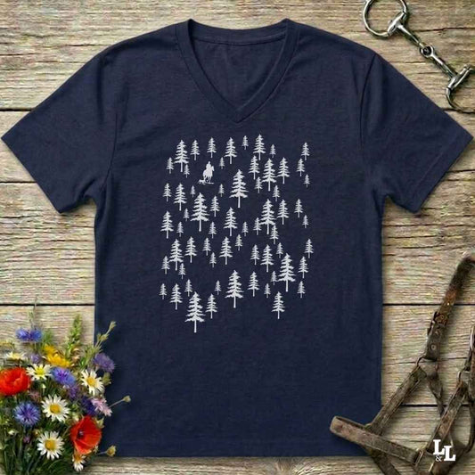 A navy blue V-neck t-shirt with a white graphic of pine trees, laid flat on a rustic wooden surface with equestrian accessories and flowers as props.