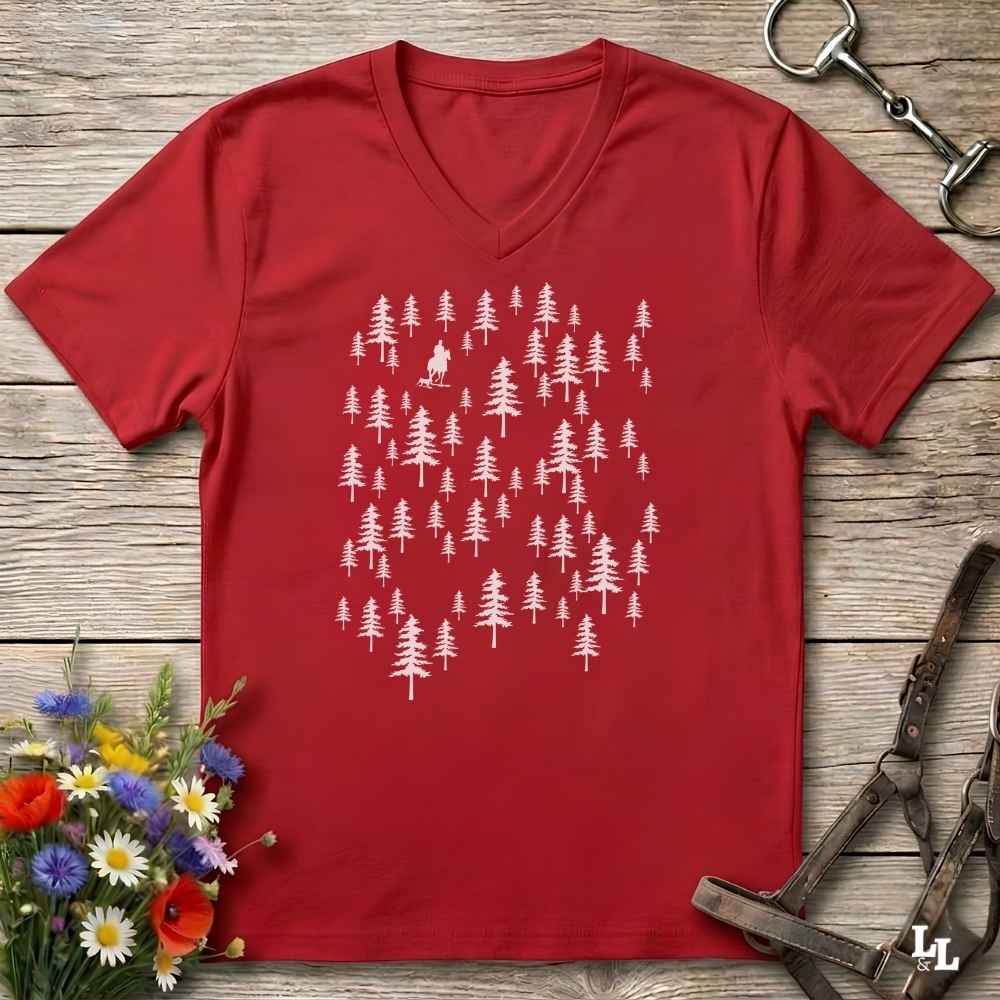 A red V-neck t-shirt with a white graphic of pine trees, laid flat on a rustic wooden background with equestrian accessories and flowers.