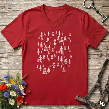 A red V-neck t-shirt with a white graphic of pine trees, laid flat on a rustic wooden background with equestrian accessories and flowers.