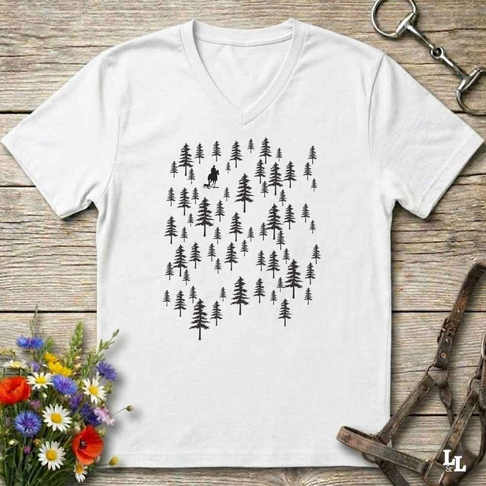 A white V-neck t-shirt with a black graphic of pine trees and a small hiker silhouette, laid flat on a rustic wooden surface with flowers and leather gear props.