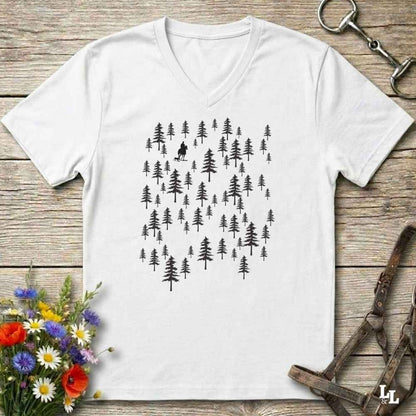 A white V-neck t-shirt with a black graphic of pine trees and a small hiker silhouette, laid flat on a rustic wooden surface with flowers and leather gear props.