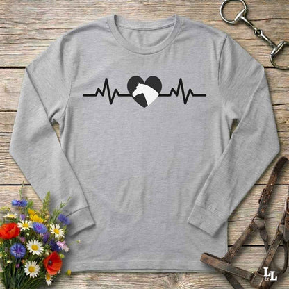 A light heather gray long-sleeve t-shirt with a black and white horse heartbeat graphic on the chest, displayed in a flat-lay arrangement on a rustic wooden background with flowers and leather tack.