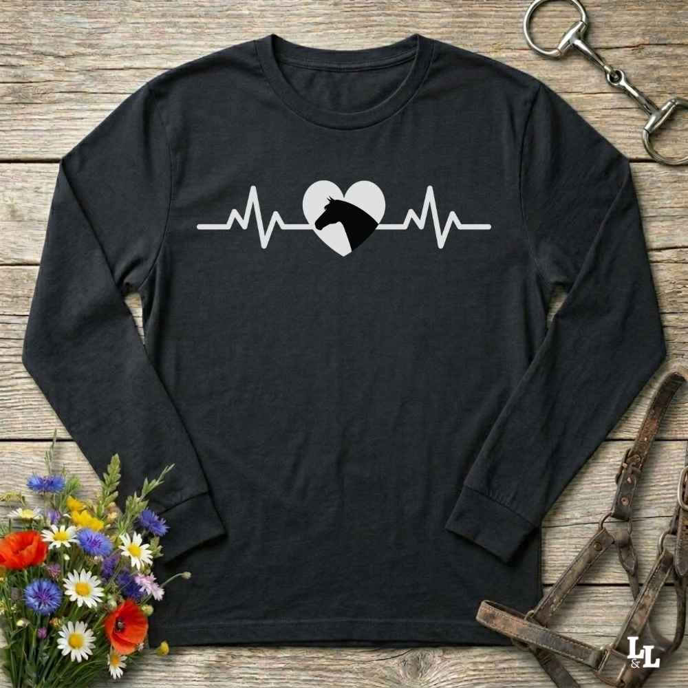 A black long-sleeve crewneck t-shirt with a white and black heartbeat graphic featuring a horse head inside a heart, displayed on a rustic wooden background with decorative flowers and horse tack.