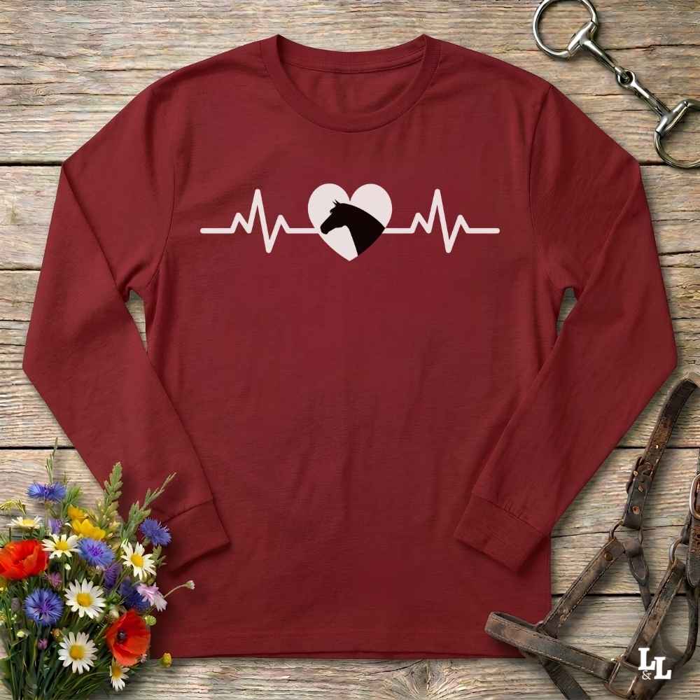 A long-sleeve crewneck shirt in maroon red, featuring a white heartbeat line graphic with a white heart and black horse silhouette. The shirt is laid flat on a rustic wooden background, surrounded by equestrian-themed props including a horse bit, leather reins, and a small bouquet of wildflowers.