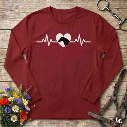 A long-sleeve crewneck shirt in maroon red, featuring a white heartbeat line graphic with a white heart and black horse silhouette. The shirt is laid flat on a rustic wooden background, surrounded by equestrian-themed props including a horse bit, leather reins, and a small bouquet of wildflowers.
