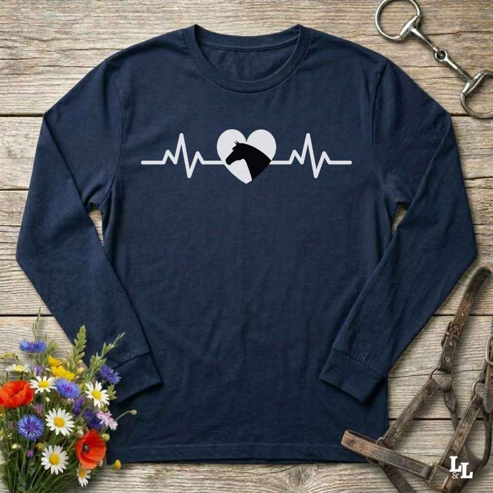 A navy blue long-sleeve t-shirt with a white heartbeat line and horse head graphic on the chest, laid flat on a rustic wooden background with equestrian accessories and flowers.
