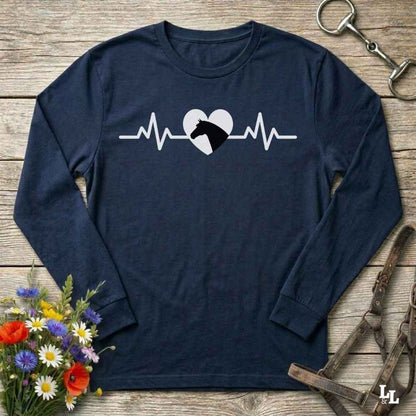 A navy blue long-sleeve t-shirt with a white heartbeat line and horse head graphic on the chest, laid flat on a rustic wooden background with equestrian accessories and flowers.