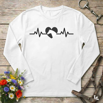 A white long-sleeve crewneck t-shirt with a black graphic of a horse heartbeat line and heart, laid flat on a rustic wooden background with equestrian accessories and flowers.