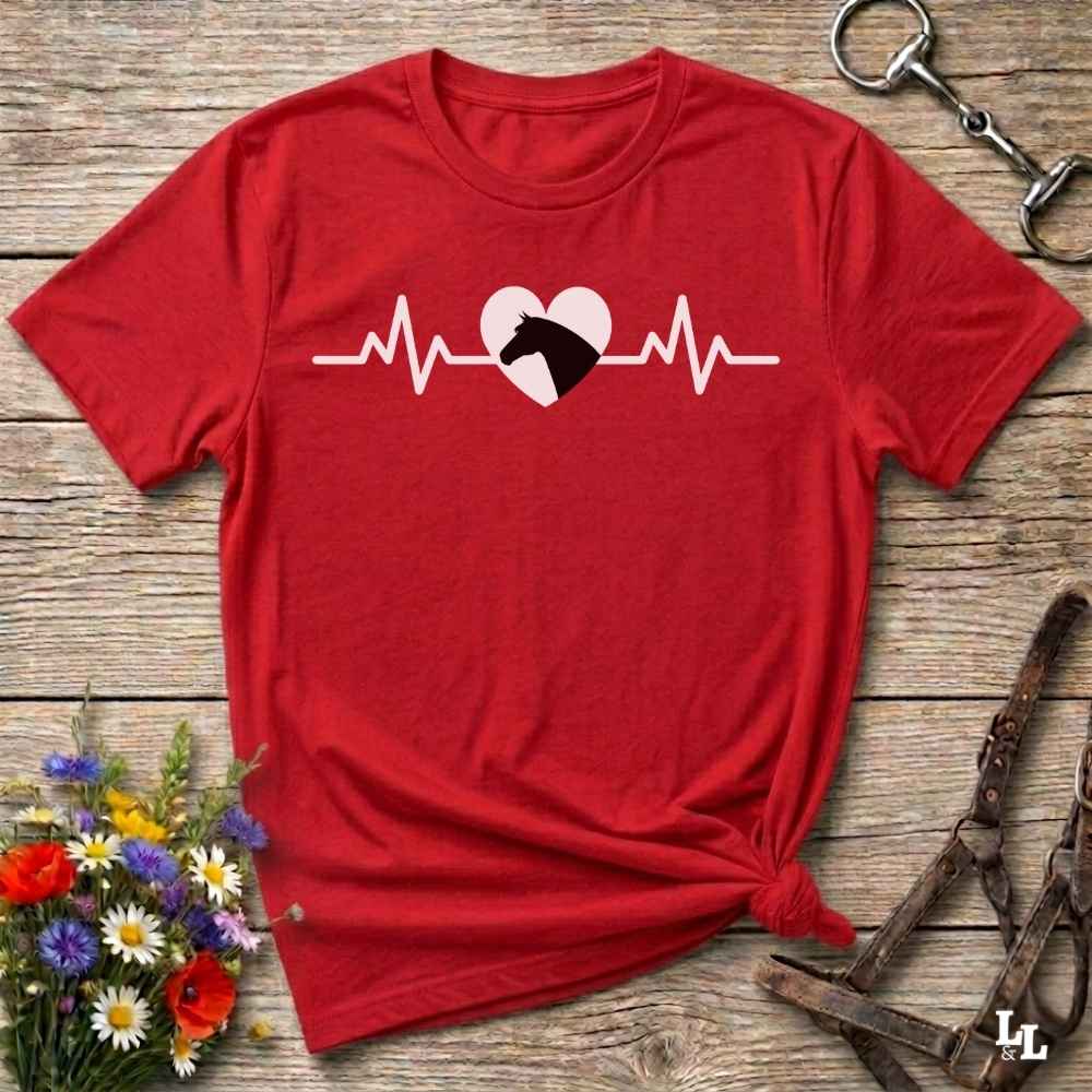 Red t-shirt with heartbeat line and horse head in the middle of the heart, on wooden background with flowers and equestrian bit and halter