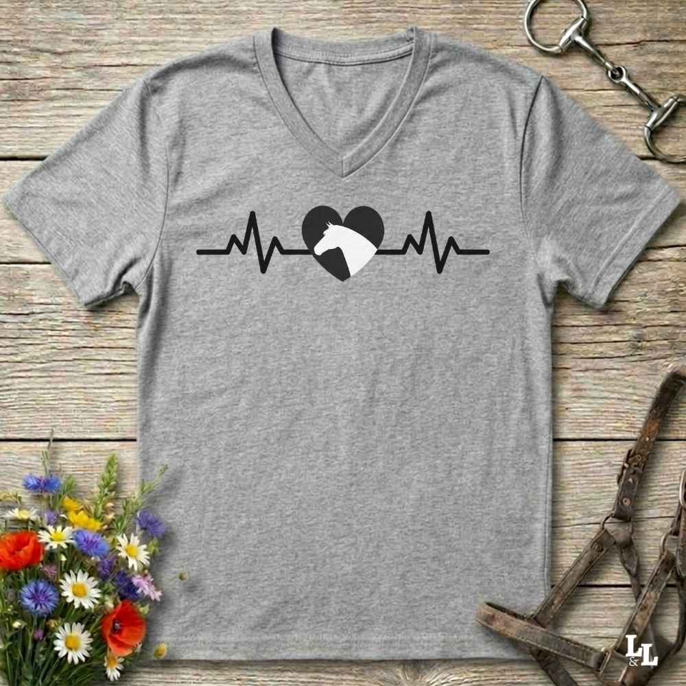 A heather gray V-neck t-shirt with a black heartbeat line and heart graphic, where the left side of the heart is black and the right side is a white horse silhouette. The shirt is displayed flat on a rustic wooden background with decorative horse tack, flowers, and a leather strap.