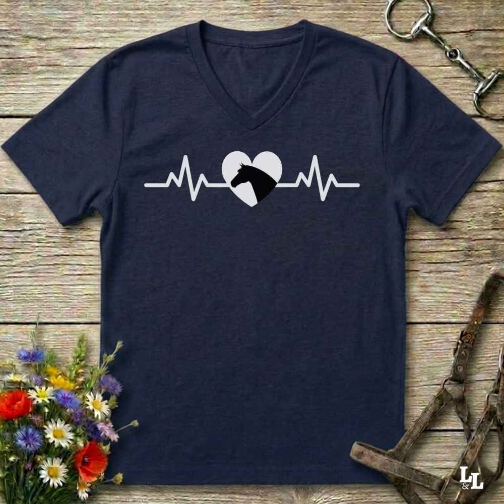 A navy blue V-neck t-shirt with a white heartbeat line graphic and a black horse head inside a white heart, laid flat on a rustic wooden surface with decorative flowers and horse tack.