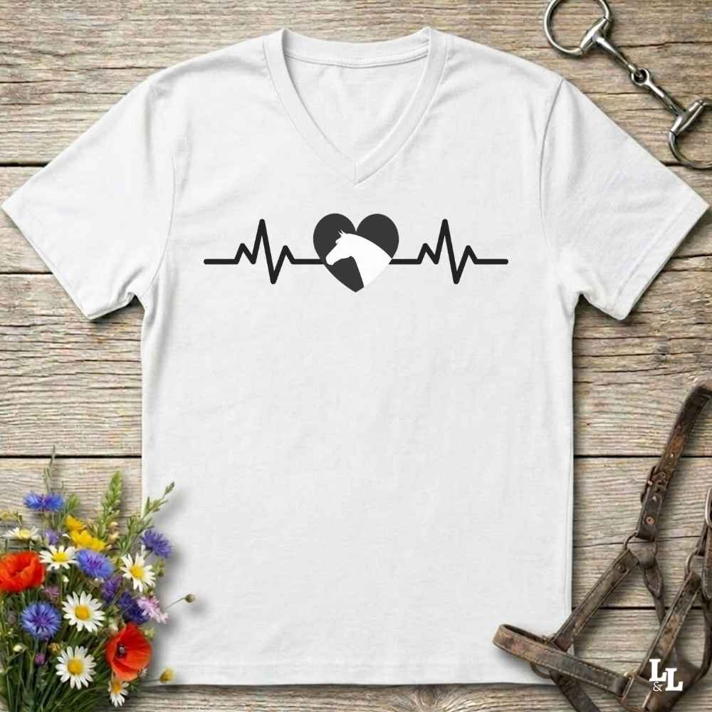 A white V-neck t-shirt with a black heartbeat line and horse silhouette graphic, laid flat on a rustic wooden background with flowers and horse tack props.