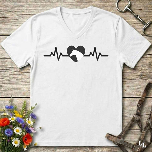 A white V-neck t-shirt with a black heartbeat line and horse silhouette graphic, laid flat on a rustic wooden background with flowers and horse tack props.