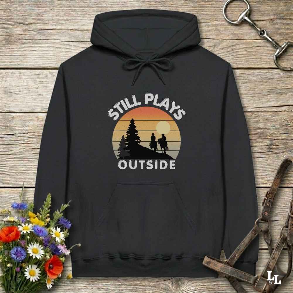 A black pullover hoodie laid flat on a rustic wood surface, featuring a vintage-style sunset graphic with the words “STILL PLAYS OUTSIDE” and silhouettes of trees and two people. Decorative flowers and horse tack are placed around the hoodie.