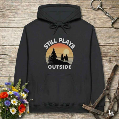 A black pullover hoodie laid flat on a rustic wood surface, featuring a vintage-style sunset graphic with the words “STILL PLAYS OUTSIDE” and silhouettes of trees and two people. Decorative flowers and horse tack are placed around the hoodie.