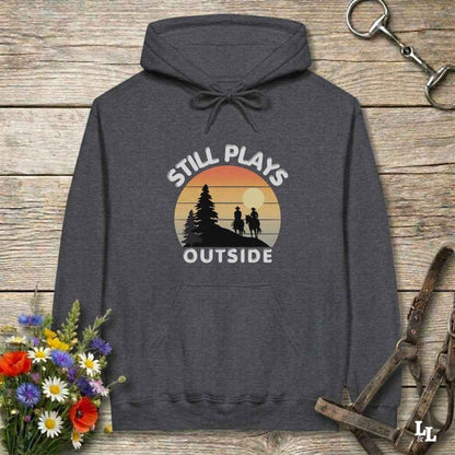 A dark gray hoodie laid flat on a rustic wooden surface, featuring a retro sunset graphic with the words 'STILL PLAYS OUTSIDE' and a silhouette of two people hiking. The hoodie is styled with flowers, a horse bit, and a leather bridle around it.
