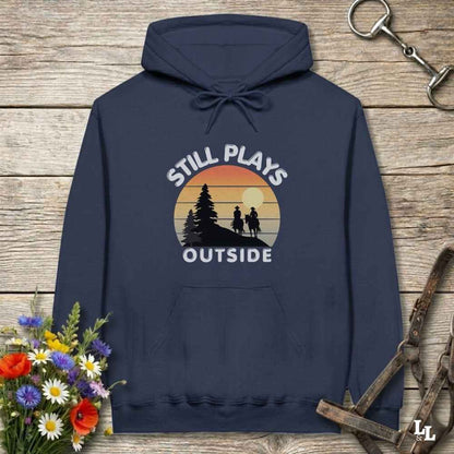 A navy blue hoodie laid flat on a rustic wooden surface, featuring a retro sunset graphic with the text 'STILL PLAYS OUTSIDE' and silhouettes of people hiking and a horse rider. Decorative flowers and horse tack are placed around the hoodie.