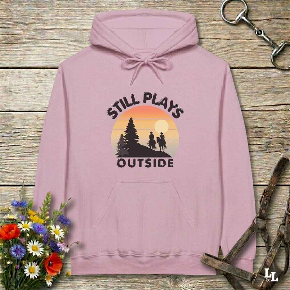 A dusty pink pullover hoodie laid flat on a rustic wooden surface, featuring a graphic print of two hikers in front of a sunset with the text 'STILL PLAYS OUTSIDE'. The hoodie is styled with a bouquet of wildflowers on the bottom left and horse tack accessories on the top right.