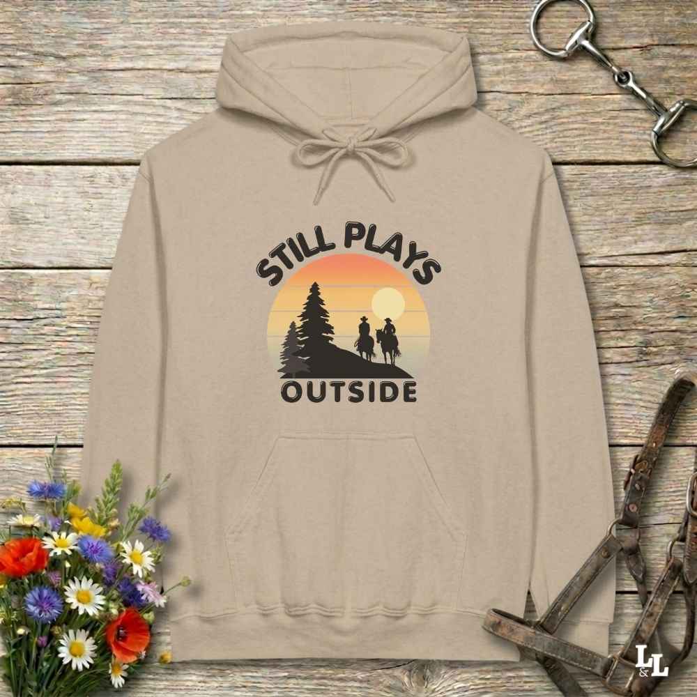 A beige pullover hoodie laid flat on a rustic wood surface, featuring a retro sunset graphic with the words 'STILL PLAYS OUTSIDE' and silhouettes of trees and two people hiking. Decorative flowers and horse tack are placed around the hoodie.