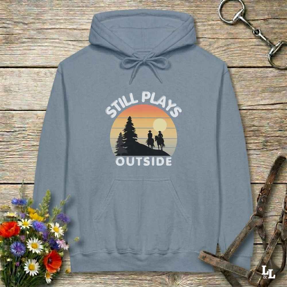A slate blue pullover hoodie with a retro sunset graphic and the text 'STILL PLAYS OUTSIDE' on the chest, displayed on a rustic wooden background with flowers and horse tack props.