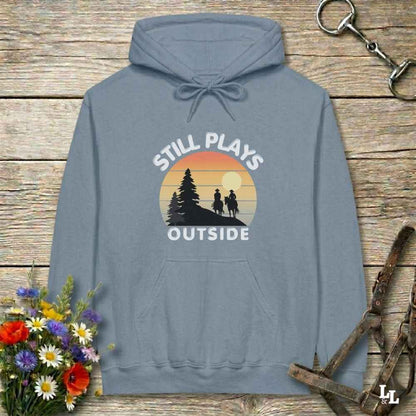 A slate blue pullover hoodie with a retro sunset graphic and the text 'STILL PLAYS OUTSIDE' on the chest, displayed on a rustic wooden background with flowers and horse tack props.
