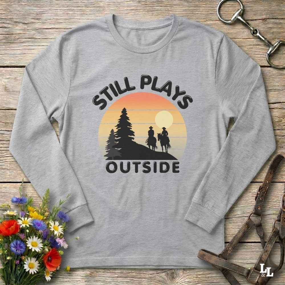 A light heather gray long-sleeve t-shirt with a retro-style graphic print. The graphic features the text 'STILL PLAYS OUTSIDE' in black arched lettering surrounding a sunset illustration with silhouettes of a cowboy and cowgirl on horseback and pine trees.