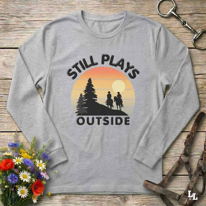 A light heather gray long-sleeve t-shirt with a retro-style graphic print. The graphic features the text 'STILL PLAYS OUTSIDE' in black arched lettering surrounding a sunset illustration with silhouettes of a cowboy and cowgirl on horseback and pine trees.
