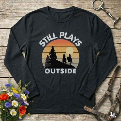 A black long-sleeve t-shirt with a retro sunset graphic and the text 'STILL PLAYS OUTSIDE' on the front, displayed on a rustic wooden background with equestrian accessories and flowers.