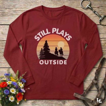 A long-sleeve crewneck t-shirt in maroon red, laid flat on a rustic wooden surface. The shirt features a retro-style graphic with the text 'STILL PLAYS OUTSIDE' surrounding a sunset silhouette of a cowboy riding a horse next to a pine tree.